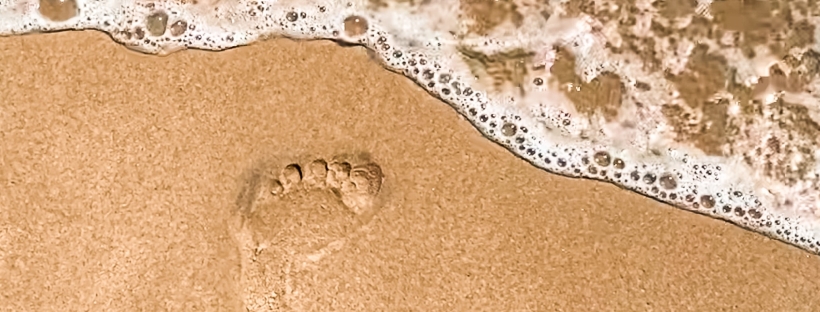 Photo of sea foam on the beach with two footprints in the sand.