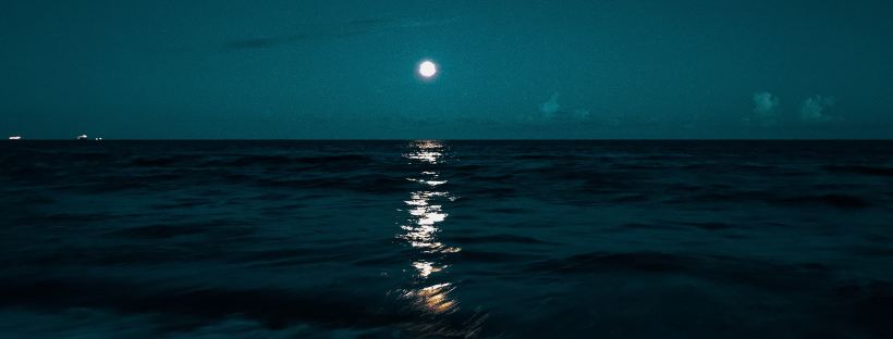 Photo of a beach waterfront at night, with dark waves and a full moon reflecting on the water.