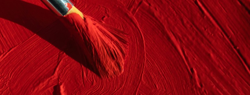 Photo of a paint brush painting on a surface with thick red paint.