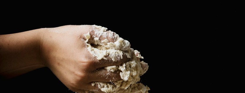 Darkly lit photo of a person’s hands with messy bread dough between their palms and fingers. A glass bowl rests under their hands on a table.
