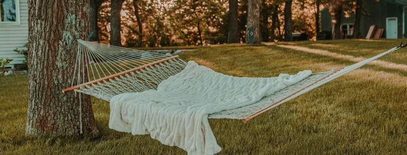 Photo of a white hammock covered in a white blanket, secured between trees. There are glimpses of a house, shed, trees and dappled light in the background.
